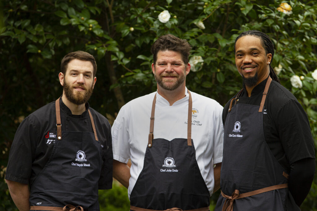 The three 2022 Chef Ambassadors stand together against a greenery background. From left to right are Chef Haydn Shaak, Chef John Ondo, and Chef Chris Williams.